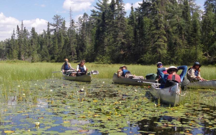 a group of teens rest in the canoes on calm water. The water is covered with lily pads, and there is a line of evergreen trees in the background. 
